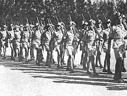Soldiers in colonial-era military uniform march with rifles shouldered.