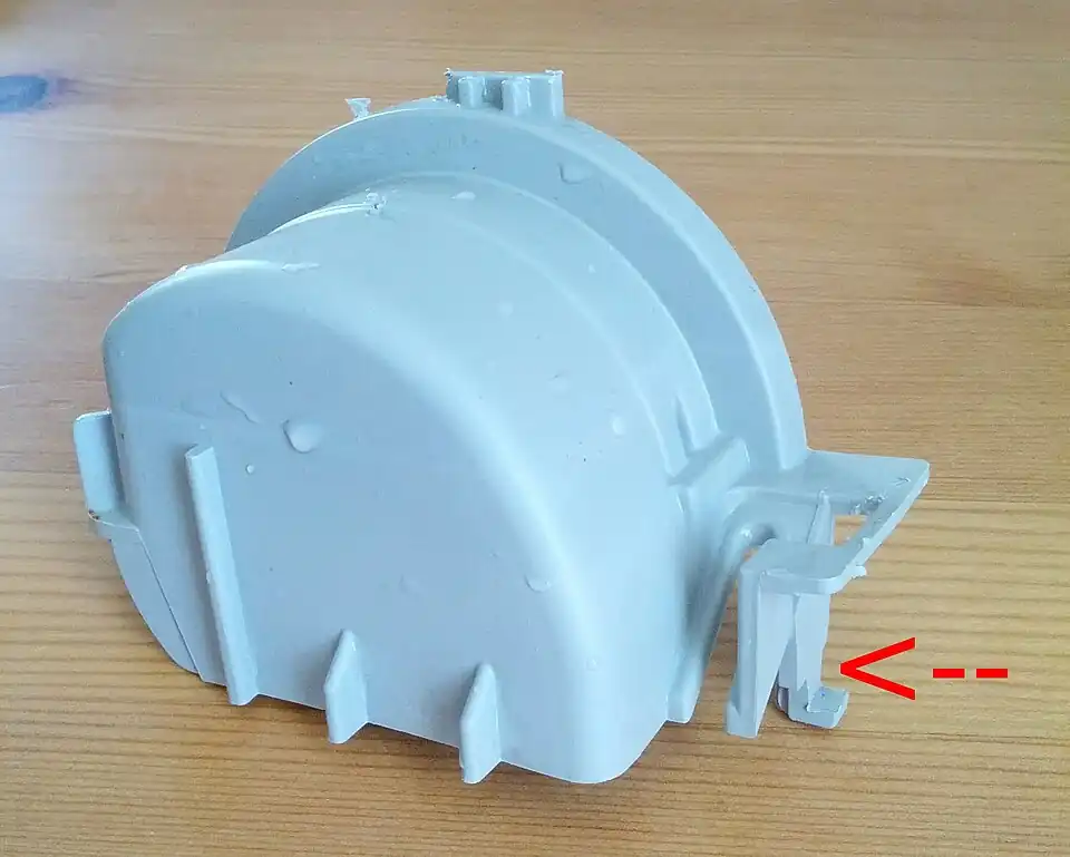 A photo of the chopper cover from a Kitchenaid kuds01fkss1. The red arrow points to a tab which prevents it from being lifted directly up -- you must either press that tab left with a screwdriver from above or pull from the left hand side by grabbing the screw hole (obscured on the left) with pliers.