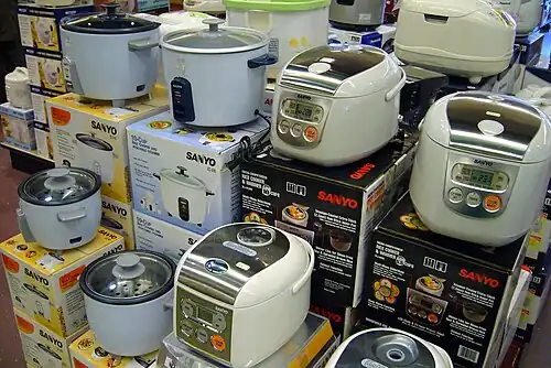 Several rice cookers on display at a store, some with one switch, others with many buttons and a digital display, each sitting atop a stack of boxes.