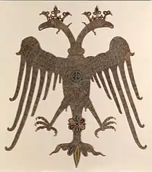 Photograph of a golden embroidered-double-headed eagle on an off-white background, with crowned heads and spread wings and legs, carrying a round medallion with Greek inscriptions on its breast.