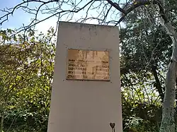 A rectangular concrete memorial stele stands outdoors, surrounded by lush trees with green and yellow leaves. Mounted on the front is a golden-yellow plaque with a small crest at the top, followed by lines of text in Georgian script and dates. Thin, bare branches hang in the foreground, framing the shot against a bright sky.