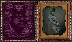 An open portrait case. On the left side, there is curved ornamentation on purple velvet. On the right, a black-and-white almost full daguerreotype portrait of a white man whittling a stick looking to the side in a top hat, tails and a striped pant.