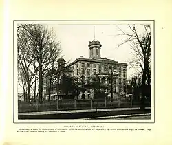 A black and white photo with a yellowing background, showing the original building as it appeared circa 1904. The building is neoclassical in style, smaller than the current one, with Ionic columns framing the entrance, a tower in the middle, an iron fence encircling it, and trees all around. There is a caption reading "Indiana Institute for the Blind. Erected 1847. Is one of the old landmarks of Indianapolis. All of the common school and many of the high school branches are taught the inmates. They are also given industrial training and instruction in music"