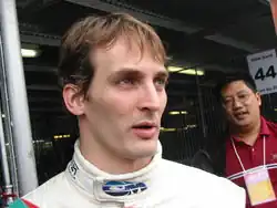 A man in his mid-twenties in white and turquoise racing overalls looking to the left of the camera