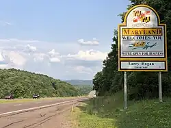 A sign adjacent to a four-lane highway reads "Maryland welcomes you. We’re Open For Business. Larry Hogan, Governor"