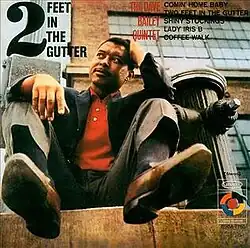 A photo of Bailey sat down on a sidewalk next to a black fire hydrant, shot from below with the soles of Bailey's shoes visible.