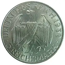 A circular silver coin, with a raised edge. Inside this is written, in raised capitals, "Graf Zeppelin/Weltflug 1929". The two halves of the inscription are separated by a depiction of the airship flying from left to right, against a background showing the northern hemisphere of the world marked with stylised lines of latitude and longitude. At the bottom of this is a letter "A".