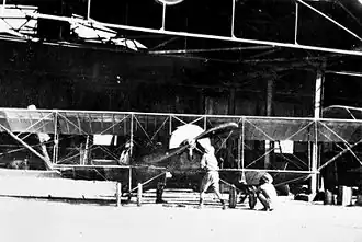 Front view of single-engined biplane and two men in front of a large open hangar