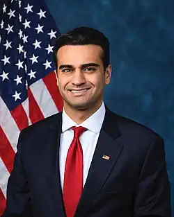 Official House portrait of Hamadeh smiling in front of the U.S. flag, wearing a black suit with American flag lapel pin, white shirt, and red tie.