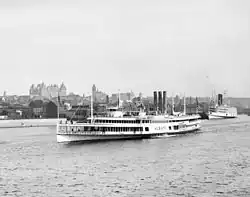 A white steam ship is seen near the shore of the Hudson River in front of the downtown area of Albany; the New York State Capitol can be seen in the background.