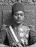 A black-and-white photograph of a man with a dark moustache wearing a turban and a dark jacket and sitting on a throne topped by two metal lions