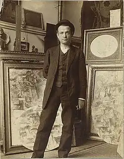 Sepia-toned photograph of a man in a suit standing in front of easels and paintings