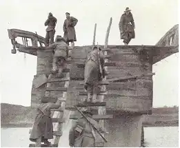 Three soldiers are standing on top of a partially destroyed bridge, while other soldiers climb up two roughly constructed ladders with their rifles slung over their shoulders while carrying stores.
