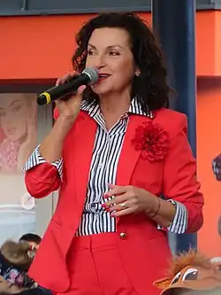 Beáta Dubasová standingwearing a blue-and-white striped shirt and a bright red pantsuit with a lapel flower, speaking or singing into a microphone amid a group of possibly seated children, and looking right of camera