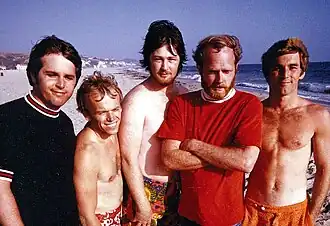 A photo of five men are standing on the beach looking at the camera. Three of the men are shirtless