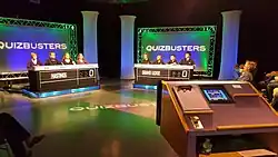 The set of a TV show titled QuizBusters. Two teams of four contestants are seated at tables that contain nameplates and a score display. Behind each is a background lit in green with the name of the show in white. Seen off to the right is the host's lectern.