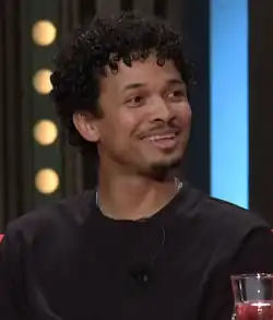 close-up of Benny Cristo wearing a black sweatshirt with a lapel microphone, grinning and looking left of camera