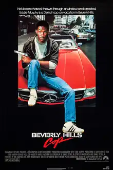 Axel Foley (portrayed by Eddie Murphy) sits on the hood of a red Mercedes-Benz convertible with a pistol pointing out on his left hand and his left foot resting above the word "Hills". The caption above reads "He's been chased, thrown through a window, and arrested. Eddie Murphy is a Detroit cop on vacation in Beverly Hills."