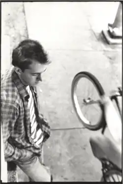 Black and white image of Bill Daniel in his 20s wearing long-sleeved plaid shirt over striped t-shirt leaning against stoop (out of frame) looking down while cyclist (his brother Lee Daniel) rides by with front tire in air. Base of streetlight in upper right corner of frame. Sidewalk scene in front of 504 W 24th Street, Austin, Texas