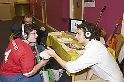 People gathered around a table wearing headphones. The journalist holds the microphone for a physically disabled person to speak