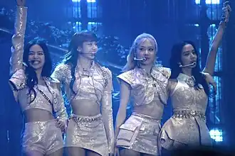Four female performers on stage, dressed in light-colored, ornate outfits. They are standing close together, smiling and posing with arms around each other or raised in celebration. They are wearing headset microphones. The stage is lit with a blue hue and background lights.