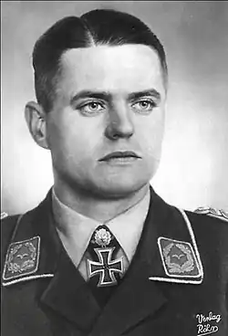 The head and shoulders of a young man, shown from the front. He wears a military uniform, with an Iron Cross displayed at the front of his white shirt collar. His facial expression is a determined and a grim smile; his eyes are looking to the left of the camera.
