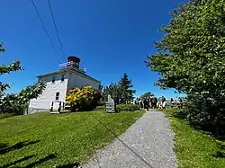 A photo taken in 2022 of the replica lighthouse (built in 1994) at Burntcoat Head Park, East Hants, Nova Scotia. The square, two story wooden lighthouse is painted white, with a hexagonal room at the top painted red, which would have housed the light to guard ship traffic in the Cobequid Bay during the age of sail. There is a group of people walking on a pathway next to the lighthouse, and trees lining the path within the public park.