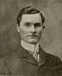 A dark-haired man in his early thirties wearing a high-collared shirt and light-colored jacket, facing left