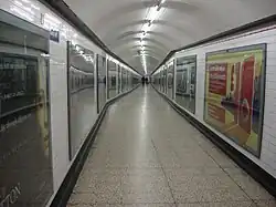 A long corridor with tiled floor and walls and a curved ceiling. The walls are lined at regular intervals with large posters framed behind glass