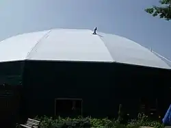 A round, green building with a white tent-like roof stands against a clear summer sky.