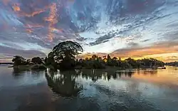 Colorful clouds and blue sky with water reflection of an island hosting a Samanea saman (rain tree) and other trees, at sunrise, in Don Det.