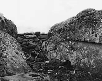 Home of a Rebel Sharpshooter - Post-battle Confederate sharpshooter body staged[12] behind the "Devil's Den barricade" illustrating "a sharpshooter…of Devil's Den" such as the one presumed killed by a percussion of a cannon shot from Little Round Top after Weed and Hazlett had been sniped.[13] Tenatively identified 2018 as a Georgia soldier.
