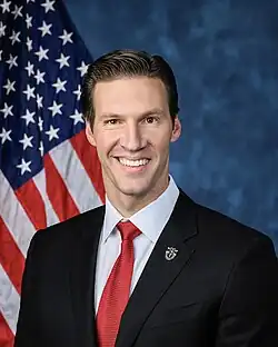 Official House portrait of Harrigan smiling in front of the U.S. flag, wearing a black suit with military medallion, white shirt, and red tie.