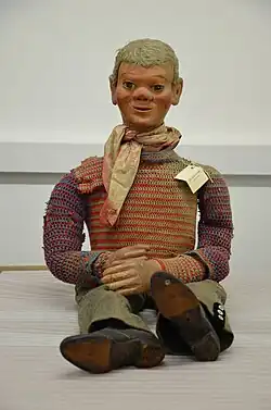 Image of male-appearing ventriloquist dummy with blond hair, faded red and cream-coloured scarf, knitted long-sleeved top in various colours, trousers, and black heeled boots, sitting seemingly unaided against a plain background.