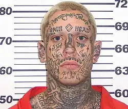Mugshot of a skinhead murderer covered in many tattoos with neo-Nazi symbolism on his face and body, including the "14" and "88" symbols on his temples.