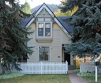 An ornate pale yellow wooden house with a pointed roofline between two tall evergreen trees. There is a white picket fence in front and a distant ridgeline in the rear.