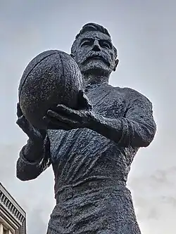 A bronze statue of a man with a large moustache, holding a rugby ball with both hands.