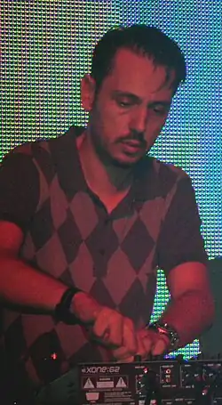 Dimitri from Paris performing on a stage with a multi-coloured background, using a machine and wearing a diamond checked shirt