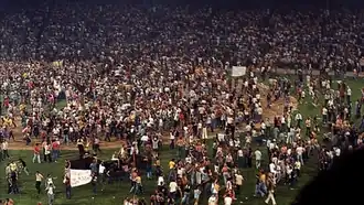 A color image of a crowd of young adults, mostly white men, walking around a baseball field and cheering. Some are holding signs, but what the signs say is unclear.