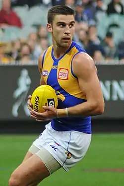 A man with brown hair holds a yellow football on a grassed playing field