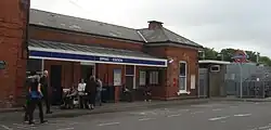 A brown-bricked building with a blue sign reading "EPPING STATION" in white letters and people in the foreground all under a blue sky with white clouds