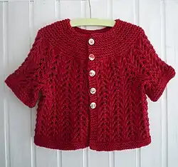 Scarlet knitted baby cardigan in lace on a clothes hanger in front of a white wall.