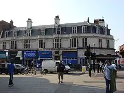 A white building with a blue-shingled roof and a blue sign reading "FINCHLEY ROAD STATION" in white letters all under a blue sky with white clouds