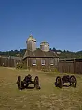 Photograph of the reconstruction of the Fort Ross Chapel, a wooden walled and roofed building with an evergreen-covered hillside behind.