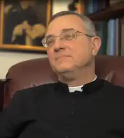A portrait photo of an older man with gray hair and glasses wearing clerical clothing sitting in a chair