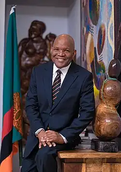 A photo portrait of Fred M'membe smiling and sitting on a desk in front of a Zambian flag and wood sculptures