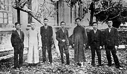 A black and white photo of seven men standing outside a building in a grassy area