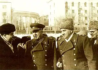 A group of men in military-style coats, with two men wearing Soviet-era uniforms, including one in a military cap and another in a fur hat. They appear to be conversing and smiling, standing outdoors in a winter setting with a background of buildings, including one with classical columns.
