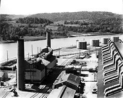 A large dark-coloured rectangular building and a smaller building with three smokestacks. In the background is the river. The steam plant is a small building with two smokestacks.