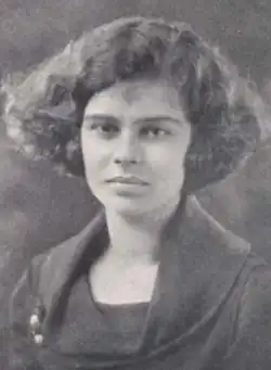 A young white woman with thick wavy bobbed hair, wearing a dark dress with a cowl neckline